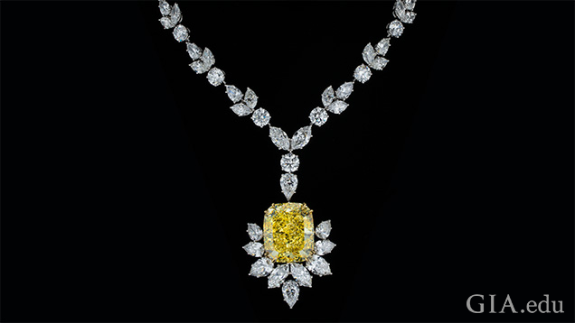 A large yellow cushion cut diamond pendant is partially framed with marquis cut diamonds and hangs from a necklace of mixed cut diamonds. A large yellow cushion cut diamond pendant is partially framed with marquis cut diamonds and hangs from a necklace of mixed cut diamonds.