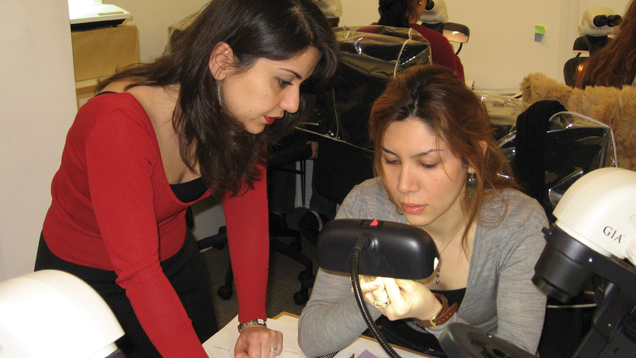 机に座って宝石を見ている女子学生の横に立つÇiğdem Lüle。