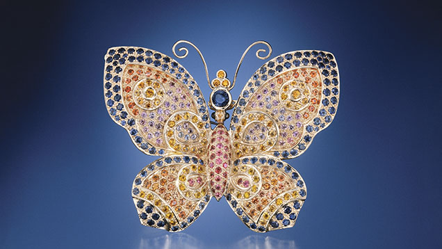 A colorful butterfly brooch adorned with various gemstones, featuring intricate designs and a shimmering finish against a blue background. A colorful butterfly brooch adorned with various gemstones, featuring intricate designs and a shimmering finish against a blue background.